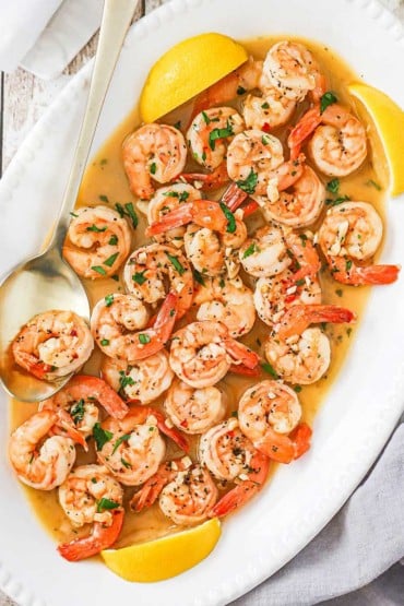 An overhead view of an oval platter filled with shrimp scampi being flanked by lemon wedges and a large golden serving spoon.