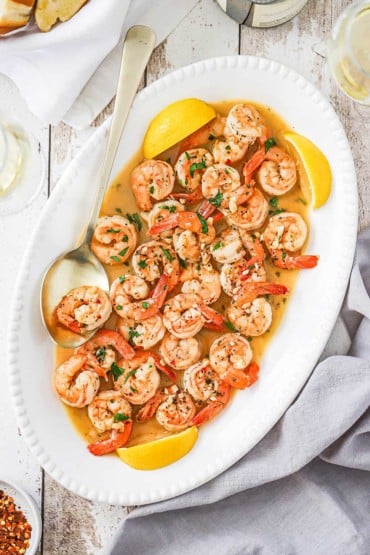 An overhead view of an oval platter filled with shrimp scampi being flanked by lemon wedges and a large golden serving spoon.