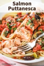 A close-up view of a salmon fillet that has been flaked apart with a fork on a bed of pasta with puttanesca sauce.