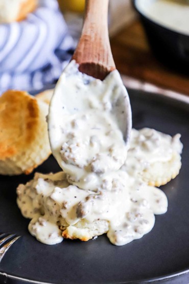 A large wooden spoon transferring sausage gravy over homemade Southern biscuits on a black plate.