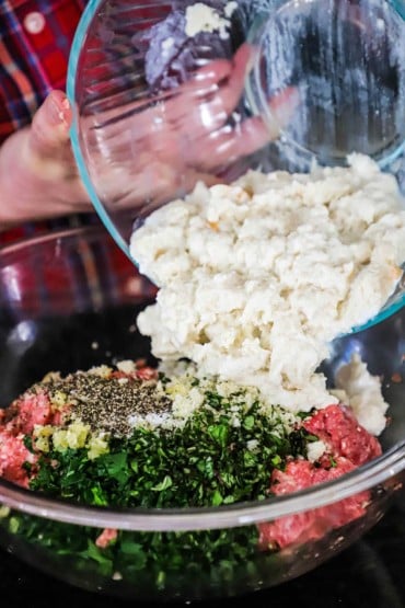 A person transferring milk-soaked bread pieces from a glass bowl into another glass bowl filled with ground meat, herbs, and seasonings.