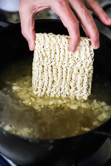 A person dropping a block of dried ramen noodles into a pot of simmering broth. 