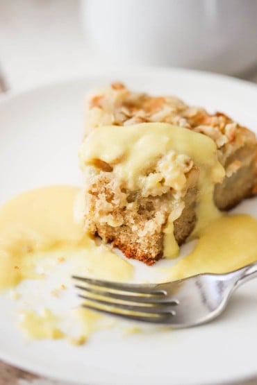 A slice of Irish apple cake with vanilla custard on a white dessert plate with a bite missing and a fork nearby.