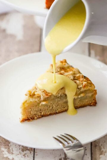 Vanilla custard being poured from a small white pitcher over a slice of Irish apple cake on a white dessert plate.