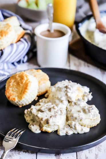 A plate filled with Southern biscuits and gravy sitting next to a white mug of black coffee and a glass of orange juice.