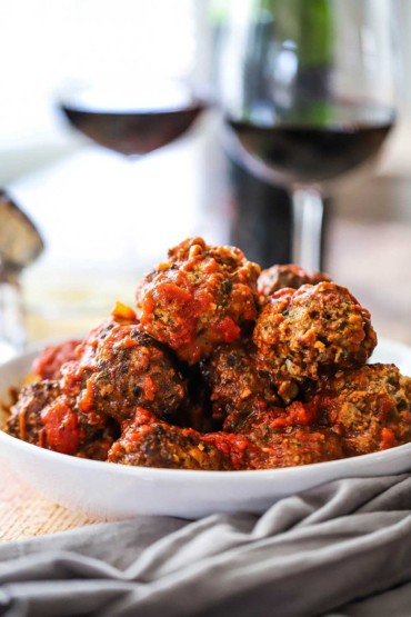 A white bowl filled with a pile of Italian meatballs sitting next to two glasses of red wine.