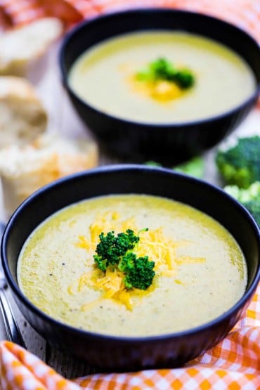 A straight-on view of two soup bowls filled with broccoli cheddar soup topped with shredded melty cheese and broccoli florets.
