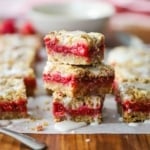 A close-up view of three raspberry Linzer bars stacked on top of each other on a sheet of parchment paper with other Linzer bars nearby.