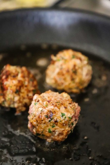 Three Italian meatballs that are being browned in a black nonstick skillet filled with a layer of olive oil.