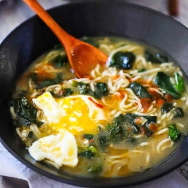 A close-up view of a black soup bowl filled with ramen with spinach and poached egg with a wooden spoon inserted into it.