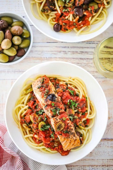 An overhead view of a bowl of salmon topped with sautéed tomatoes, olives, capers, and herbs all on a bed of pasta.