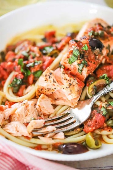 A close-up view of a salmon fillet that has been flaked apart with a fork on a bed of pasta with puttanesca sauce.