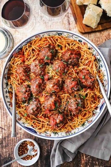 An overhead view of a large pasta bowl filled with spaghetti and meatballs with two glasses of red wine and a board of bread all nearby.