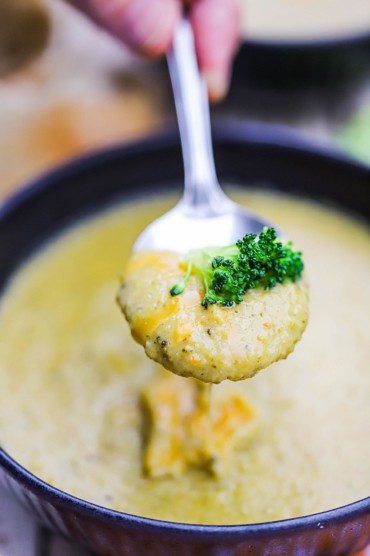 A person raising a spoon filled with broccoli cheddar soup with a broccoli floret sitting on top all over of a bowl of the soup.