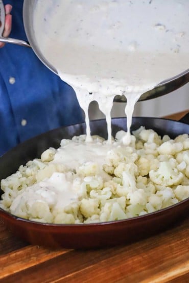 A person pouring a bechamel sauce from a large skillet into an oval baking dish filled with steamed cauliflower florets.