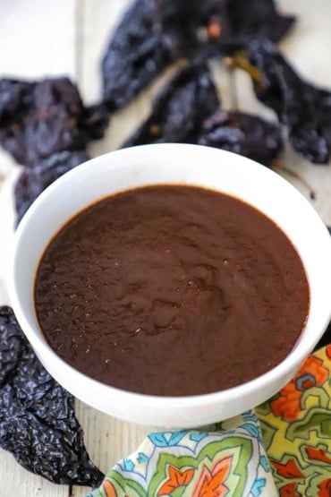 A white bowl filled a brown ancho chili sauce that is sitting next to dried ancho chilis and a festive colorful napkin.