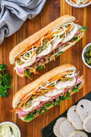 An overhead view of two fully stuffed banh mi sandwiches sitting on a cutting board with sliced cha lua, peppers, and mayonnaise nearby.