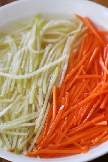 A close-up view of a white bowl filled with julienned carrots and a daikon radish that is soaking in pickling juice.