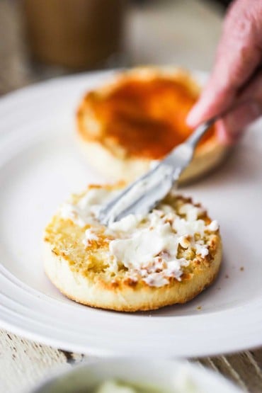 A person using a small knife to smear softened butter on an opened toasted English muffin.