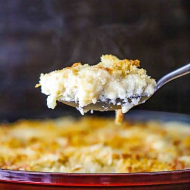 A spoon filled with cauliflower gratin is being raised over a baking dish filled with the same.