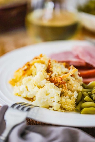 A white dinner plate filled with a serving of cauliflower gratin next to green beans and slices of cooked ham.