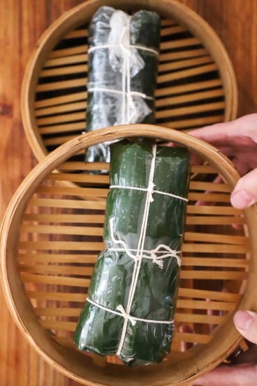 A person holding the basket of a bamboo steamer with a pork loaf that has been wrapped in banana leaves and plastic wrap and tied securely with kitchen twine.