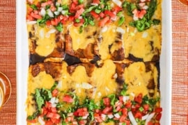 An overhead view of a white 9 by 13 baking dish filled with cheese enchiladas in ancho sauce topped with shredded lettuce and pico de Gallo.