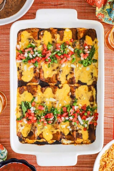 An overhead view of a white 9 by 13 baking dish filled with cheese enchiladas in ancho sauce topped with shredded lettuce and pico de Gallo.