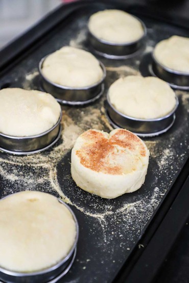 Eight uncooked English muffins on a griddle being browned with a thin layer of cornmeal.