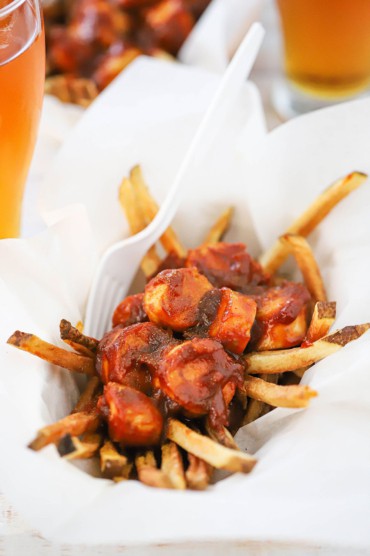 A straight-on view of a basket lined with white parchment paper that is filled with a helping of German currywurst with a plastic white fork stuck in the side.
