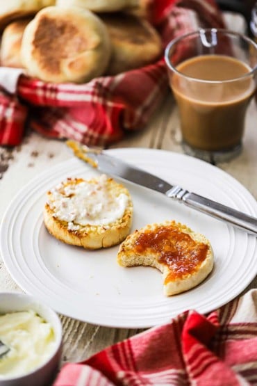Two homemade English muffin halves covered with butter and peach jam with one that has a bite taken out of it.