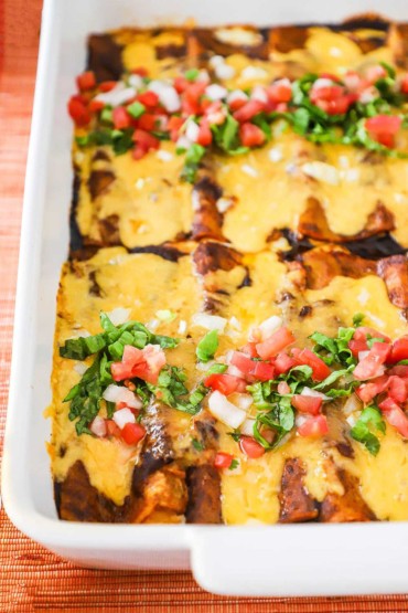 A partial and close-up view of a white baking dish filled with ancho enchiladas that are covered with melted cheese and topped with lettuce and pico de Gallo.