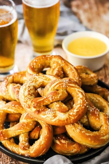 A straight-on view of a black platter filled with a stack of homemade soft pretzels next to two glasses of beer and a bowl of melted cheese.