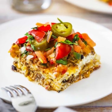 A dinner plate filled with a square serving of Mexican lasagna topped with chopped tomatoes and jalapeño slices.