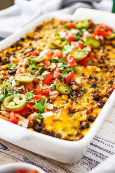 A close-up view of a Mexican lasagna in a white 9 by 13 baking dish topped with chopped tomatoes and slices of jalapeños.
