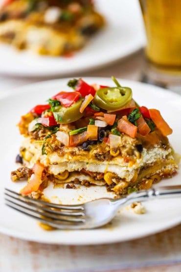 A slice of Mexican lasagna with a bite missing and a fork sitting next to it on a white dinner plate.