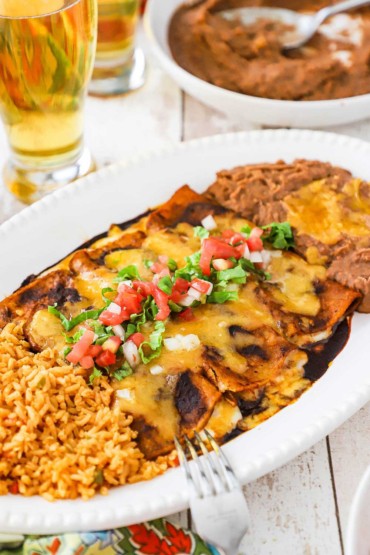A close-up view of a large white platter filled with Mexican rice on one side and refried beans on the other side and three cheese enchiladas with ancho sauce in the middle.