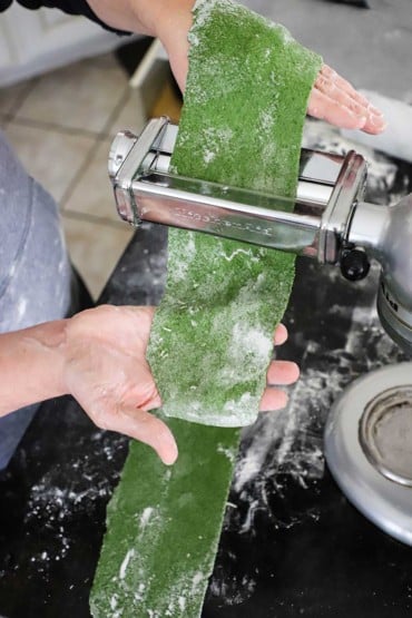 A person passing a sheet of fresh spinach pasta through a pasta attachment on a stand mixer.