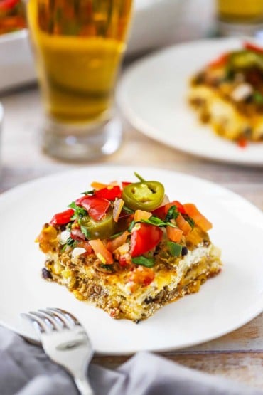 A dinner plate filled with a square serving of Mexican lasagna topped with chopped tomatoes and jalapeño slices.