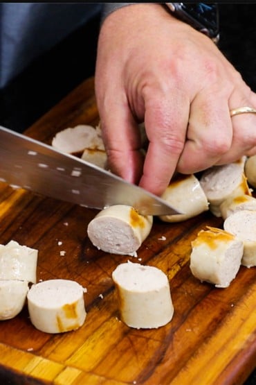A person using a large chef's knife to slice lightly grilled Bavarain bratwurst into bite-sized pieces on a small wooden cutting board.