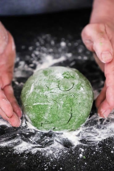 A person's hands hovering around a ball of spinach pasta sitting on a floured black marble countertop.