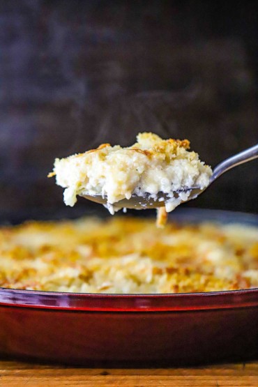 A spoon filled with cauliflower gratin is being raised over a baking dish filled with the same.