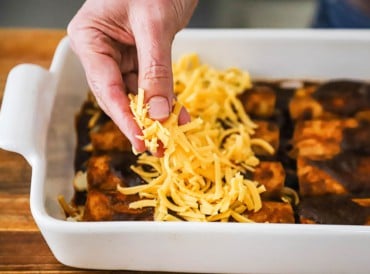 A person sprinkling shredded cheddar cheese over the tops of rolled cheese enchiladas in ancho sauce.