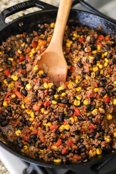 A large cast-iron skillet filled with taco meat that includes black beans, corn, and chopped tomatoes and there is a large wooden spoon inserted into the middle of it all.