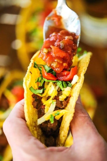 A person using one hand to hold up a crispy beef taco in one hand and drizzling salsa over the top of the taco with the other hand.