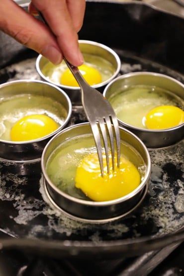 A person using a fork to bread an egg yolk open in a metal round disc that is cooking the whole egg in a skillet.