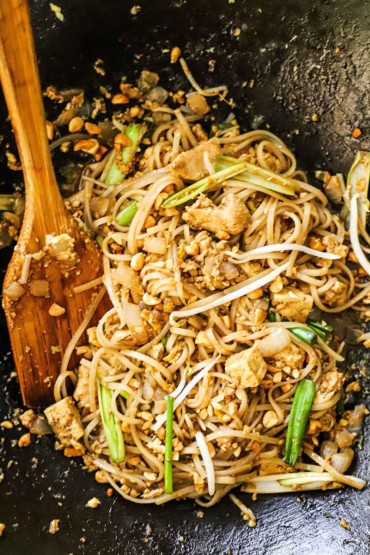 An overhead view of chicken pad Thai that has just been stir-fried in a wok with a wooden spatula off to the side.