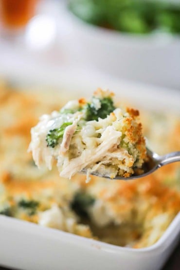 A large spoon is used to raise a helping of chicken and rice casserole with broccoli and cheese from of baking dish filled with the casserole.