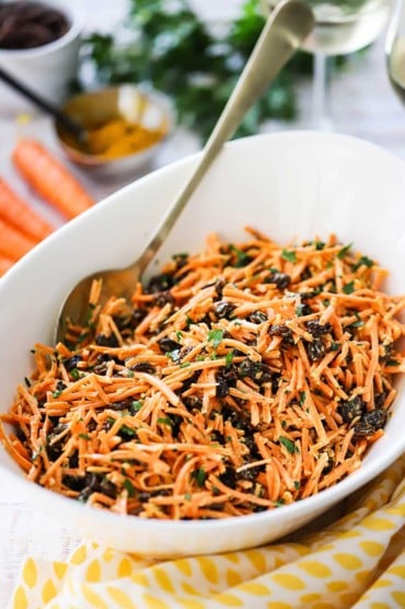 An oval white serving bowl filled with carrot and raisin salad with a gold serving spoon inserted on the side of the bowl.