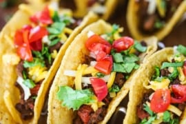 A close-up view of two rows of Tex-Mex beef tacos sitting up-right in a taco stands topped with chopped lettuce, tomatoes, and shredded cheese.